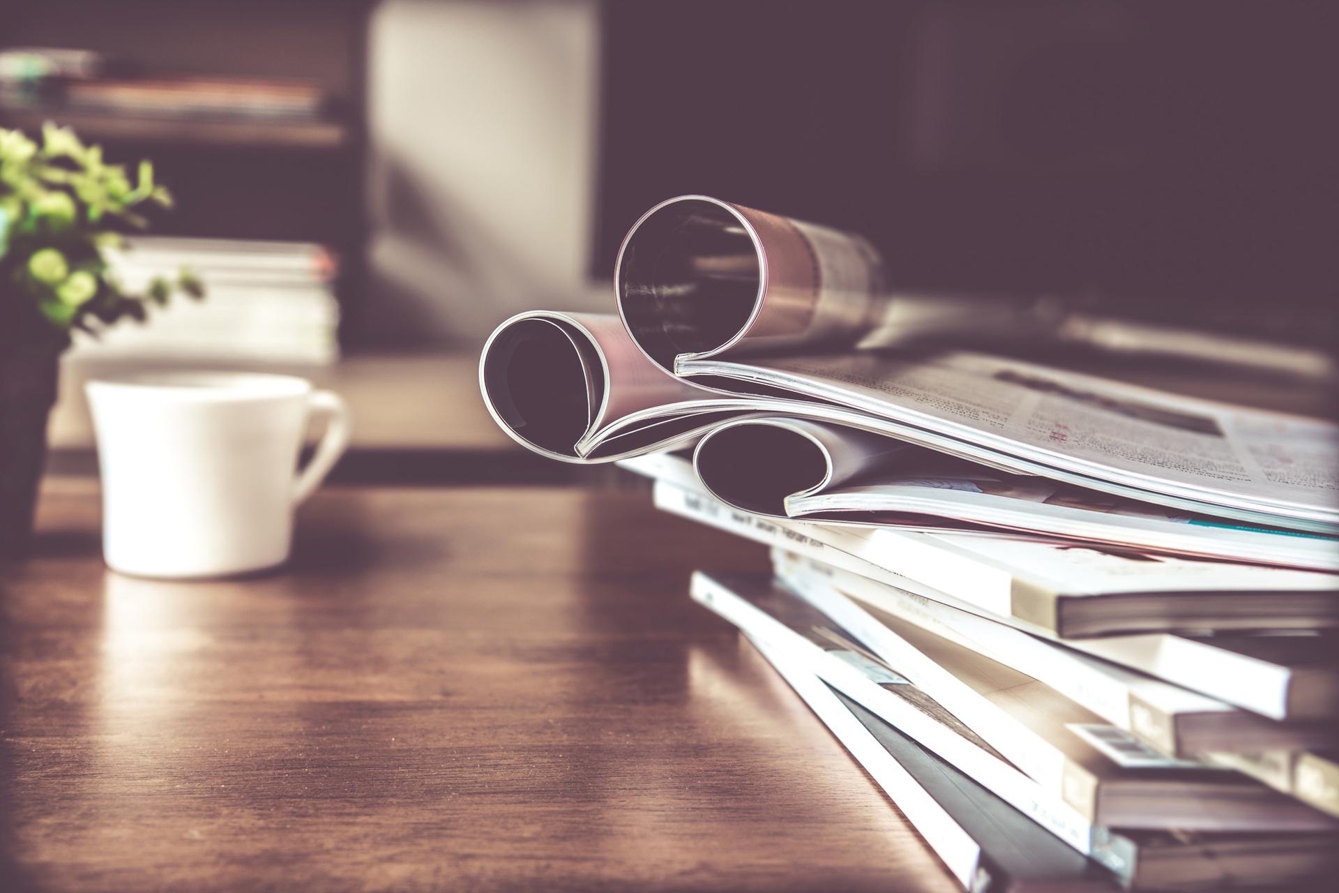 Stack of magazines with a plant and coffee cup on a wooden table