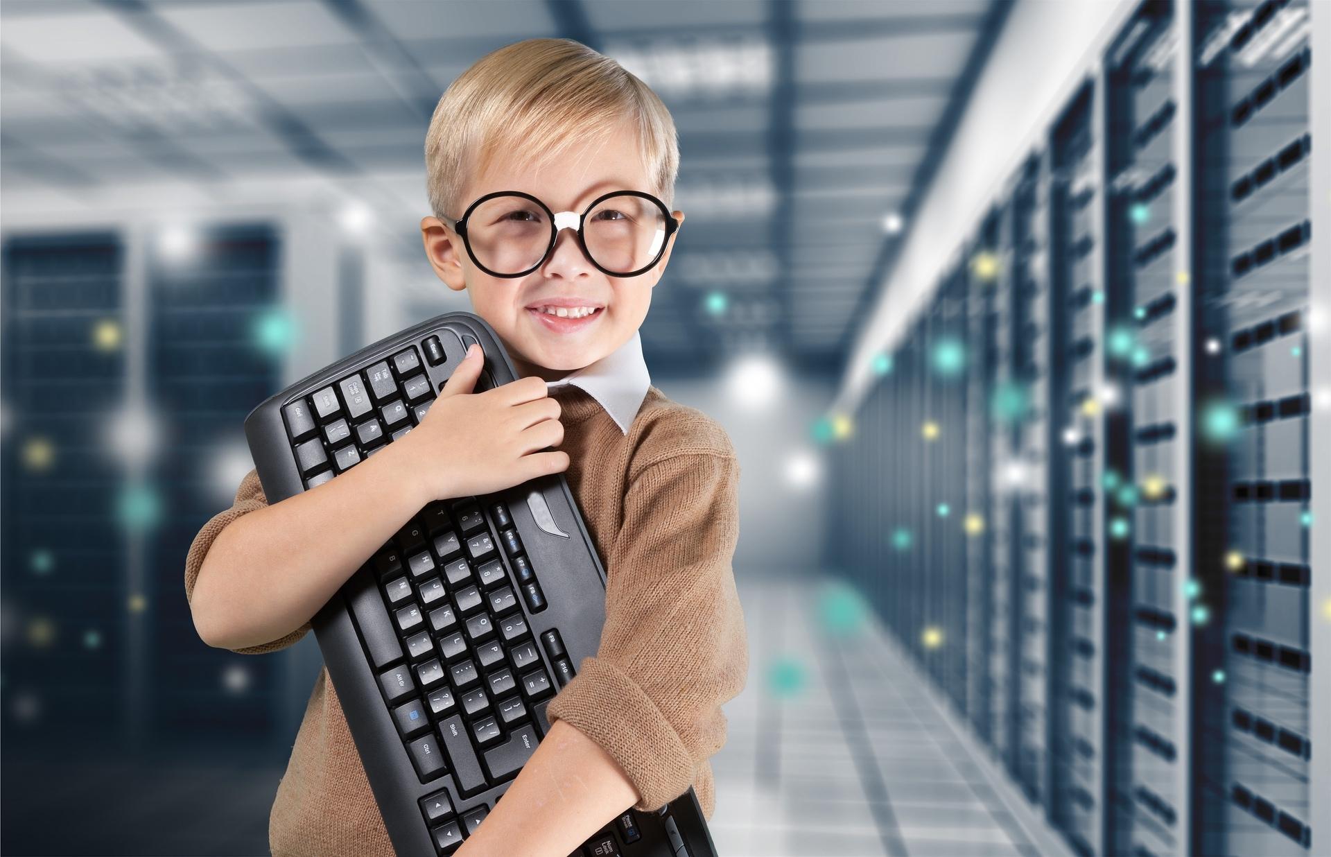 Child in glasses smiling and holding a keyboard in a server room