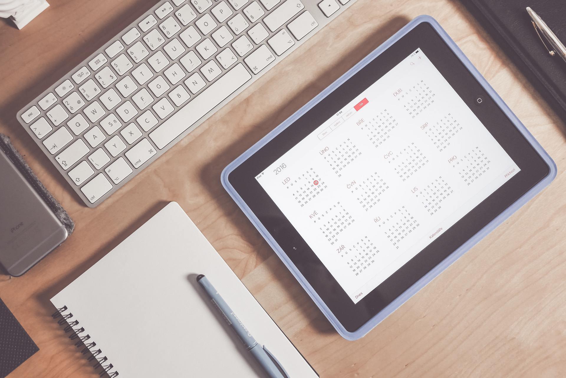 Tablet displaying a calendar on a wooden desk with a keyboard and stationery.