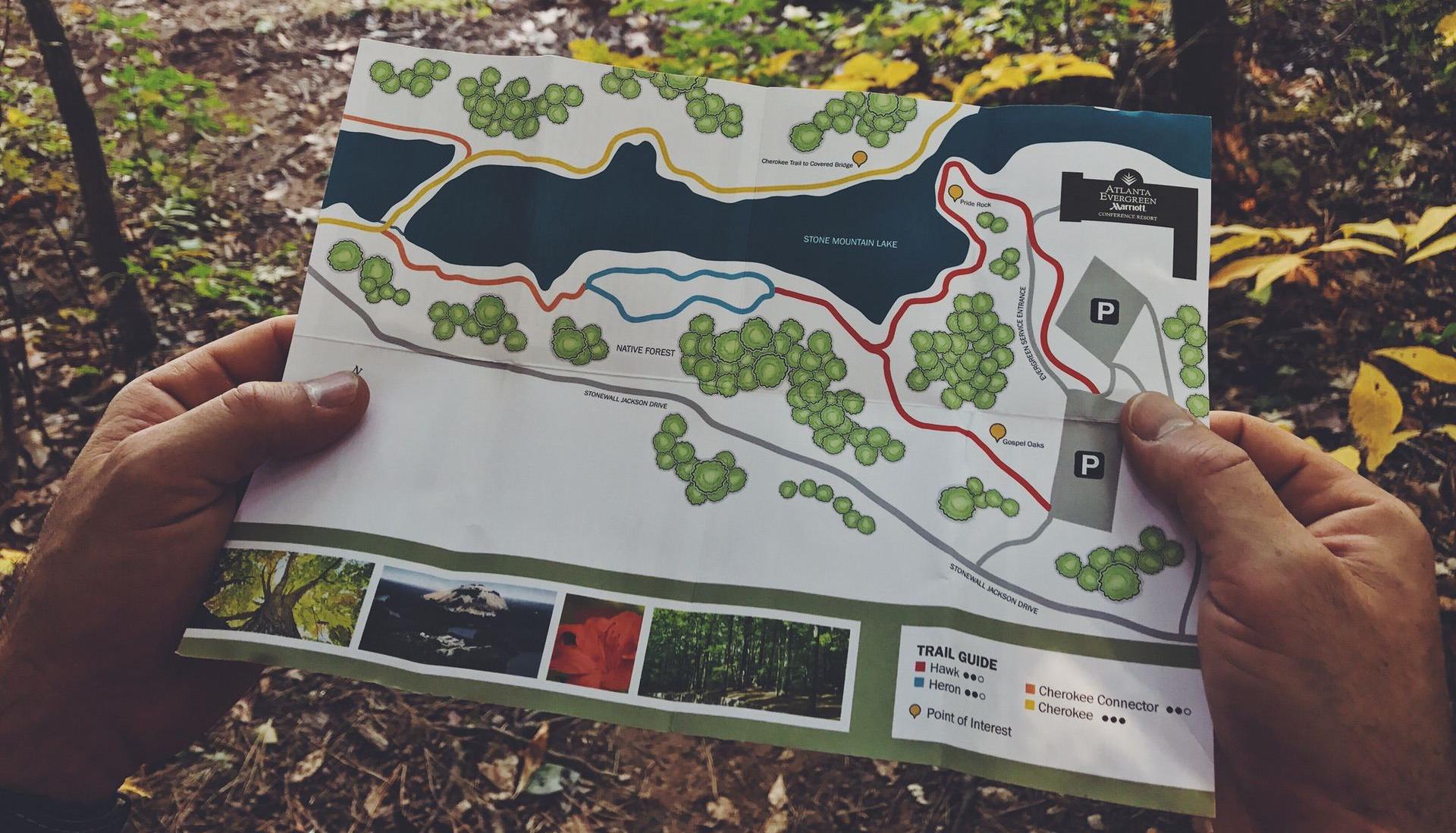 Person holding a trail map near a lake surrounded by trees and foliage