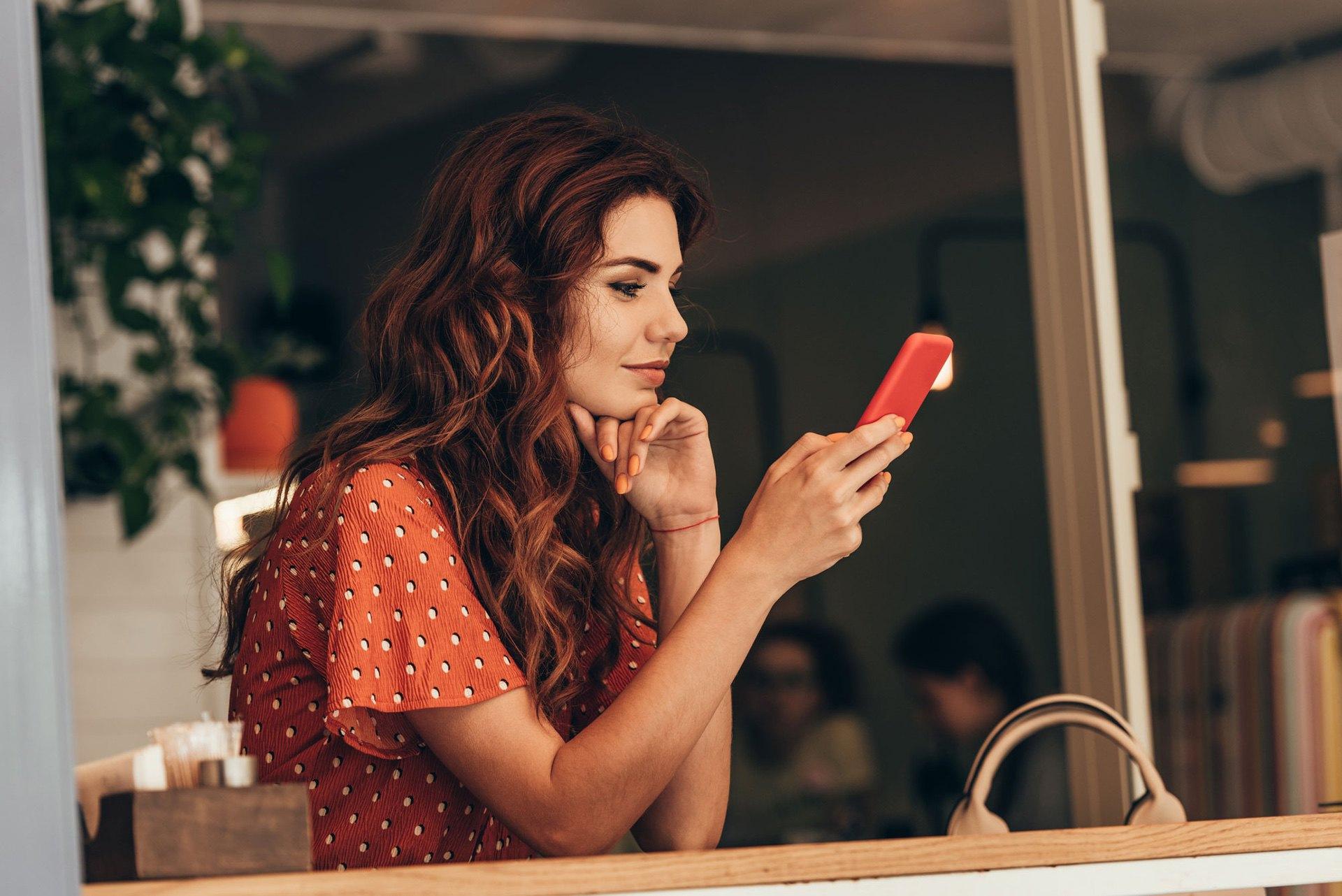 Woman with wavy hair using a red phone at a cafe, resting chin on hand