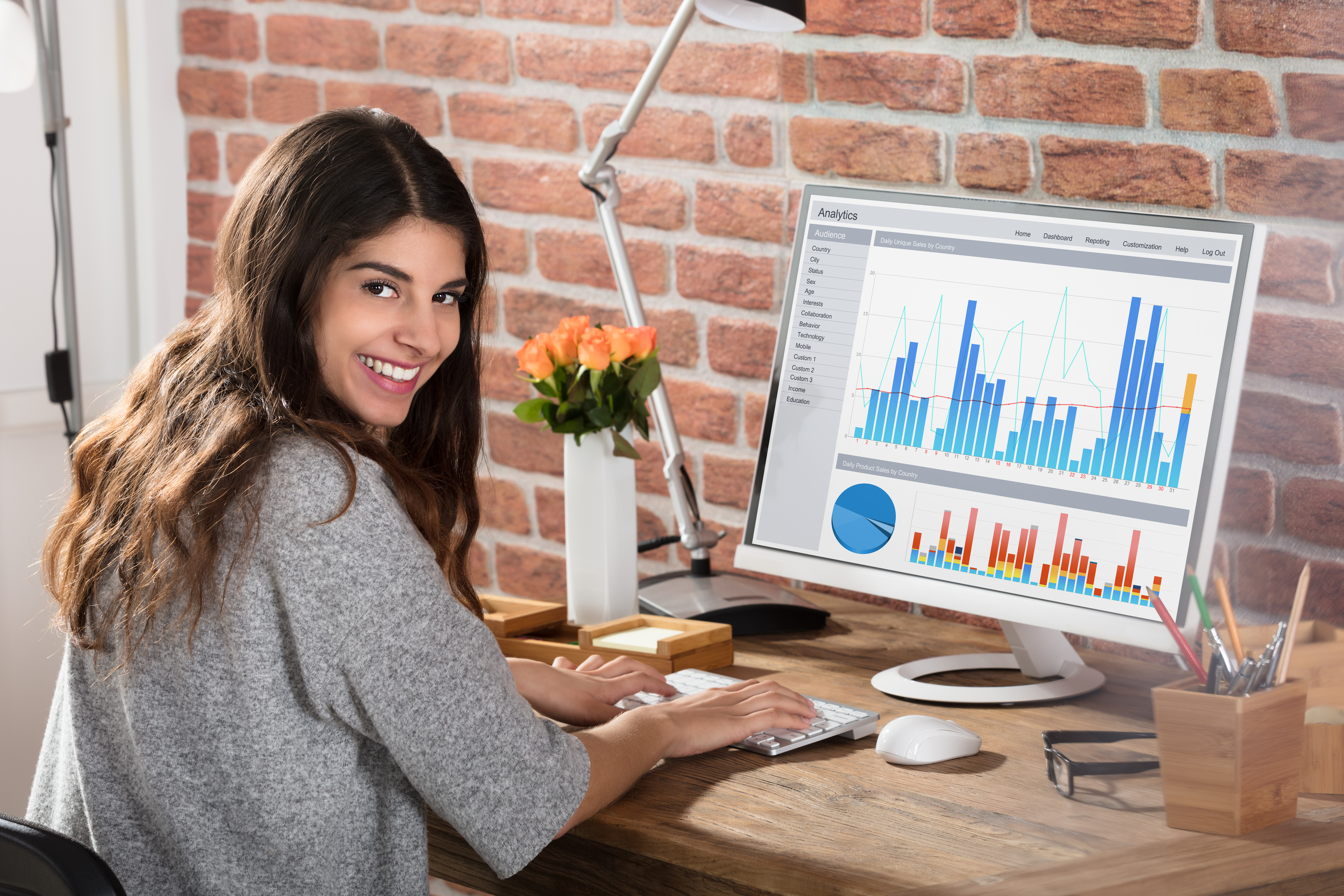 young professional woman seated at a desk in front of a monitor displaying graphs, she is turned towards the camera and smiling