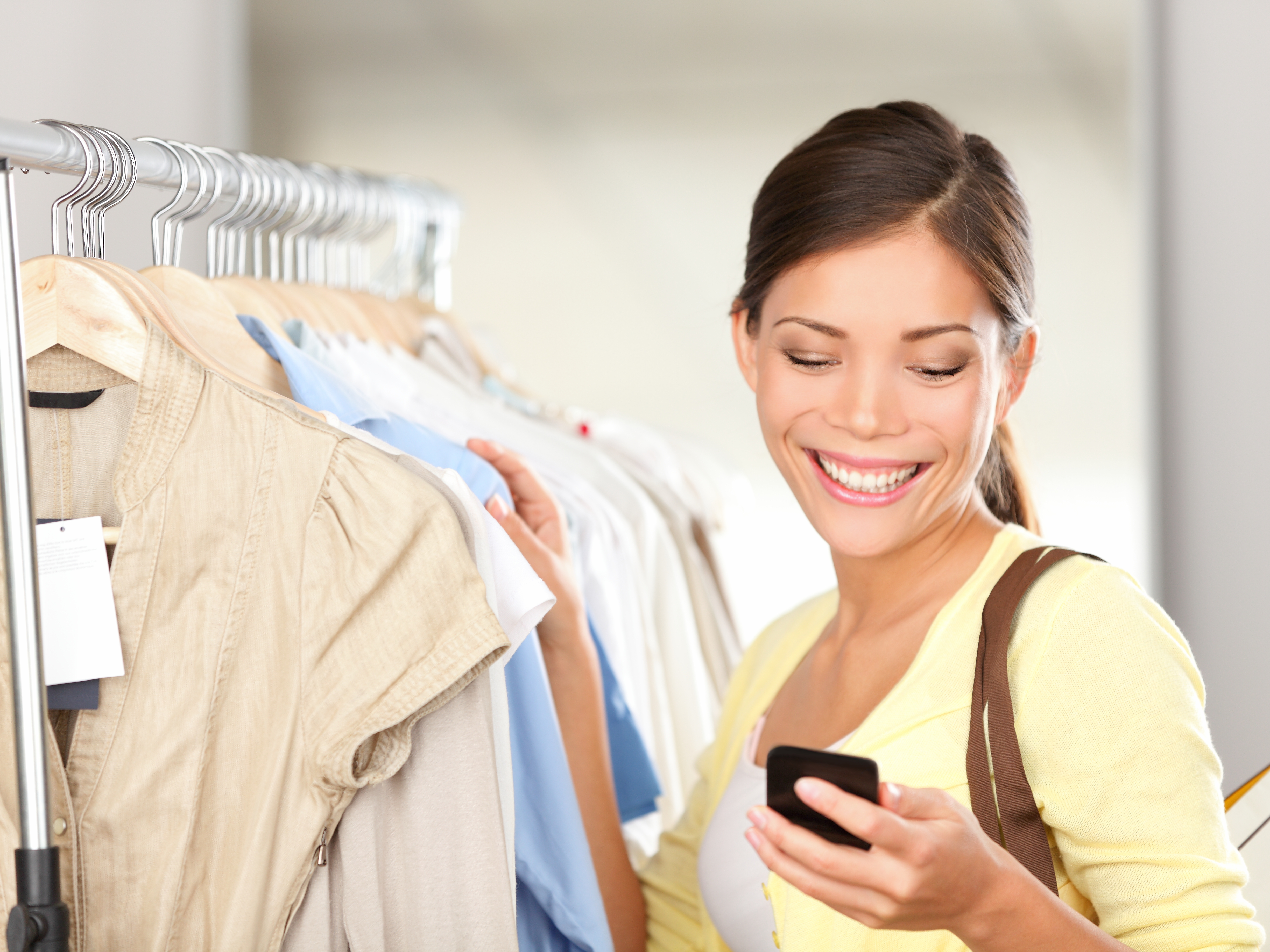 brown haired lady smiling at a phone she is holding in one hand while touching a rack of clothing with the other hand