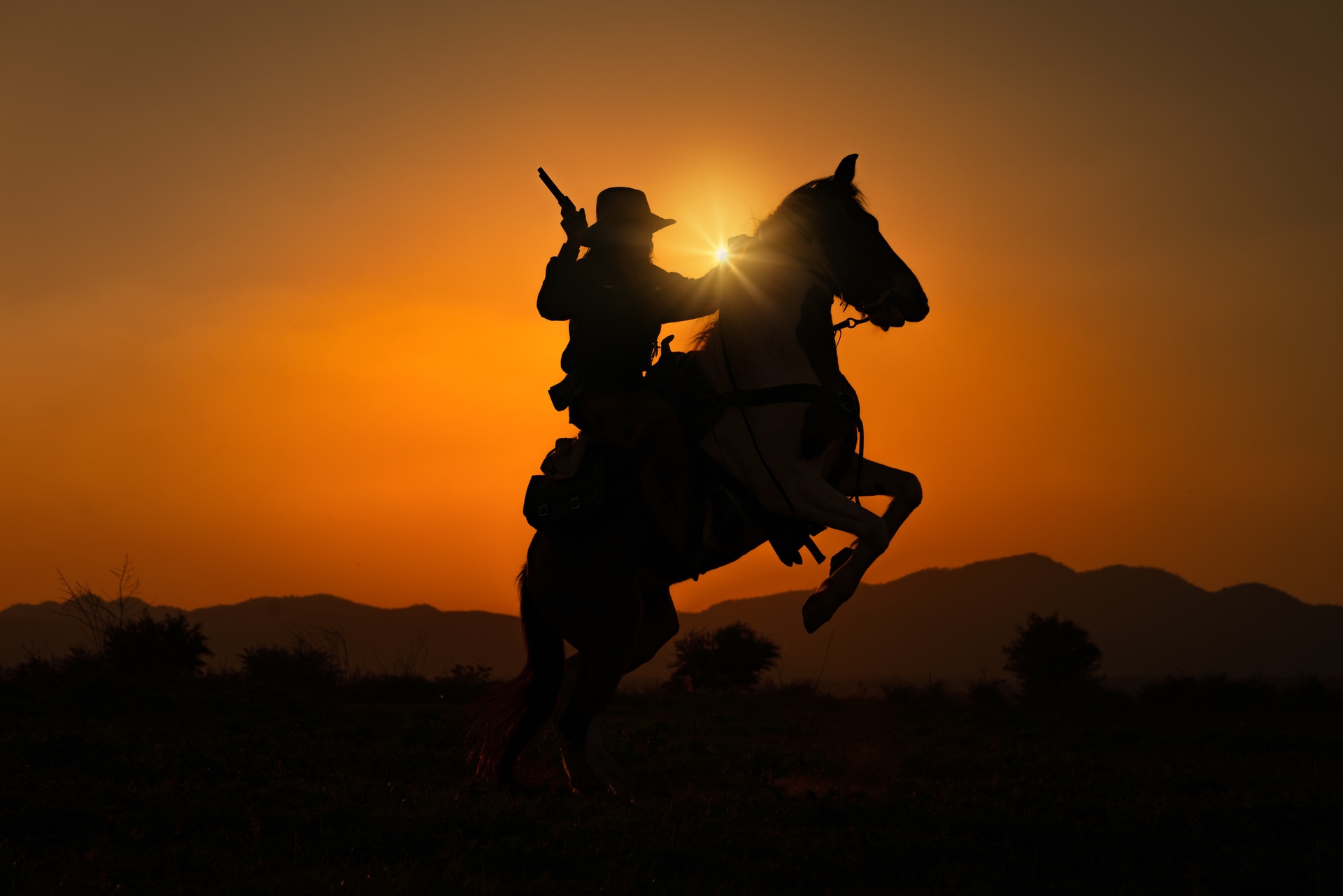 photo of a person in a brimmed hat on horseback, where the horse has its front legs up and the sun is shining behind them, making them appear as a silhouette