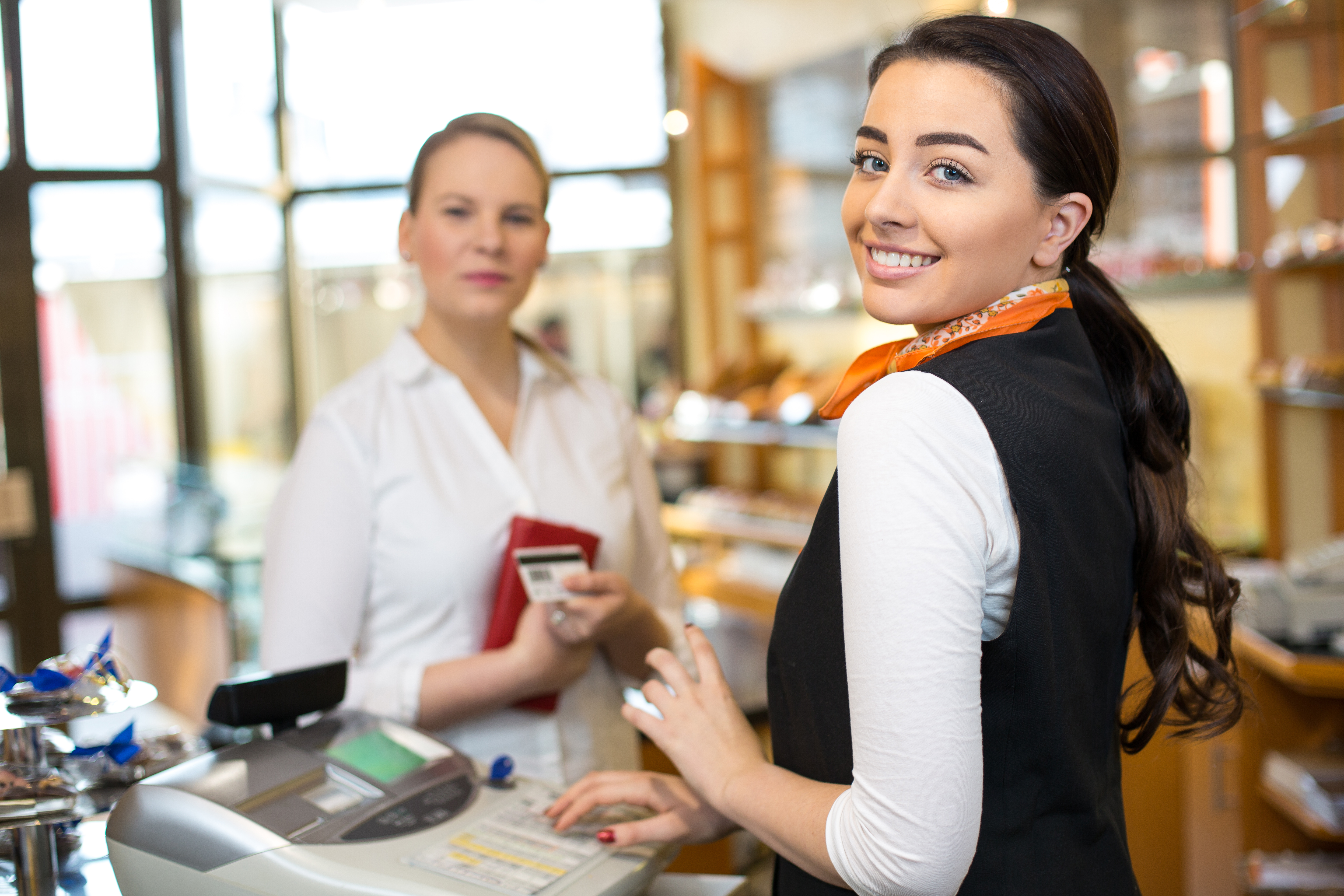 young lady holding a credit card at a cash register and smiling while another lady who is out of focus stands on the other side of a counter