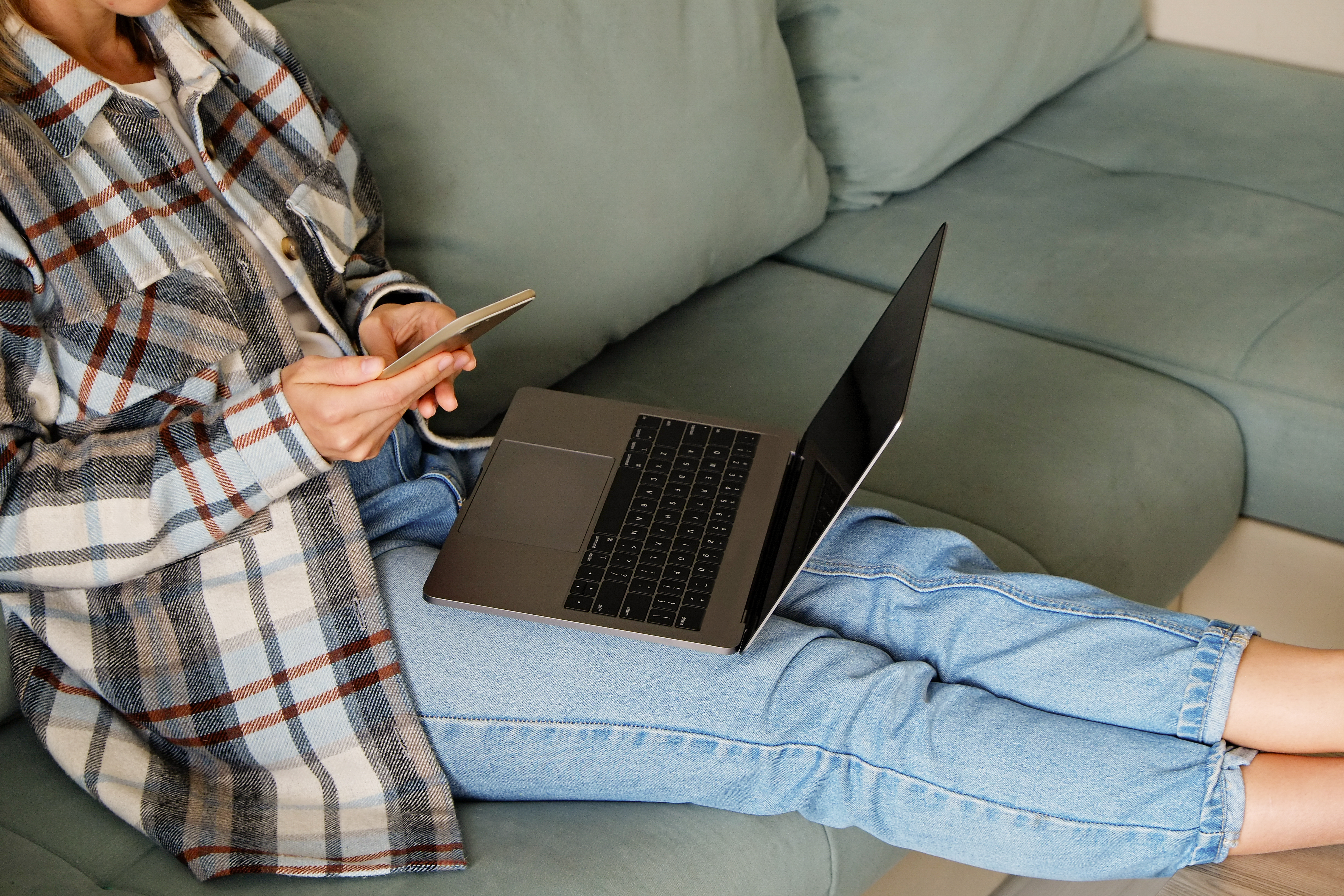 woman's body seated on a green comfy couch with her legs stretched out on a stool in front of her, she is wearing a flannel shirt and jeans and is using a cellphone and has an open laptop on her lap