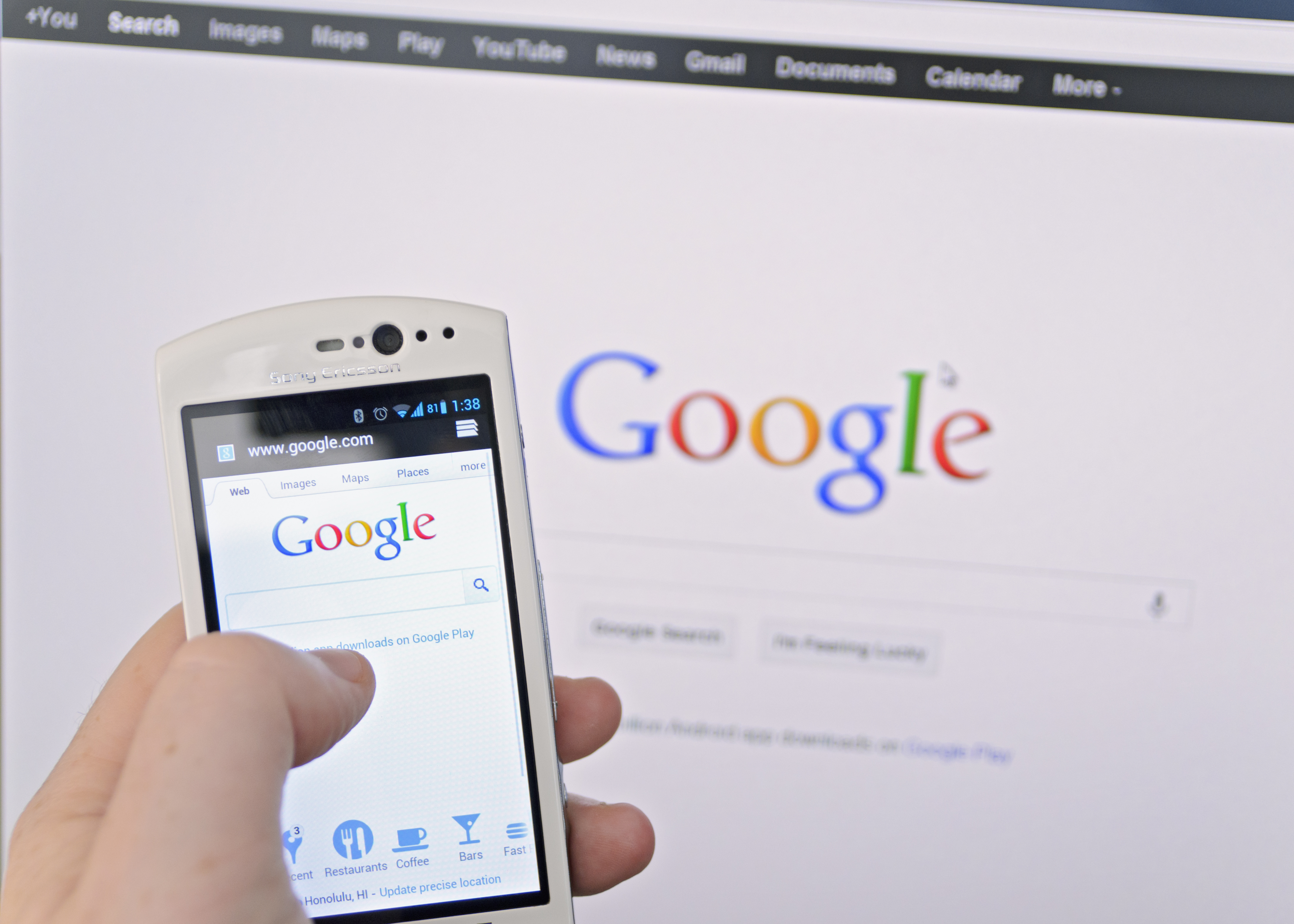 In the foreground of this picture, a hand holds a mobile phone with Google search on the screen. In the background a Google search window is open on a larger screen.