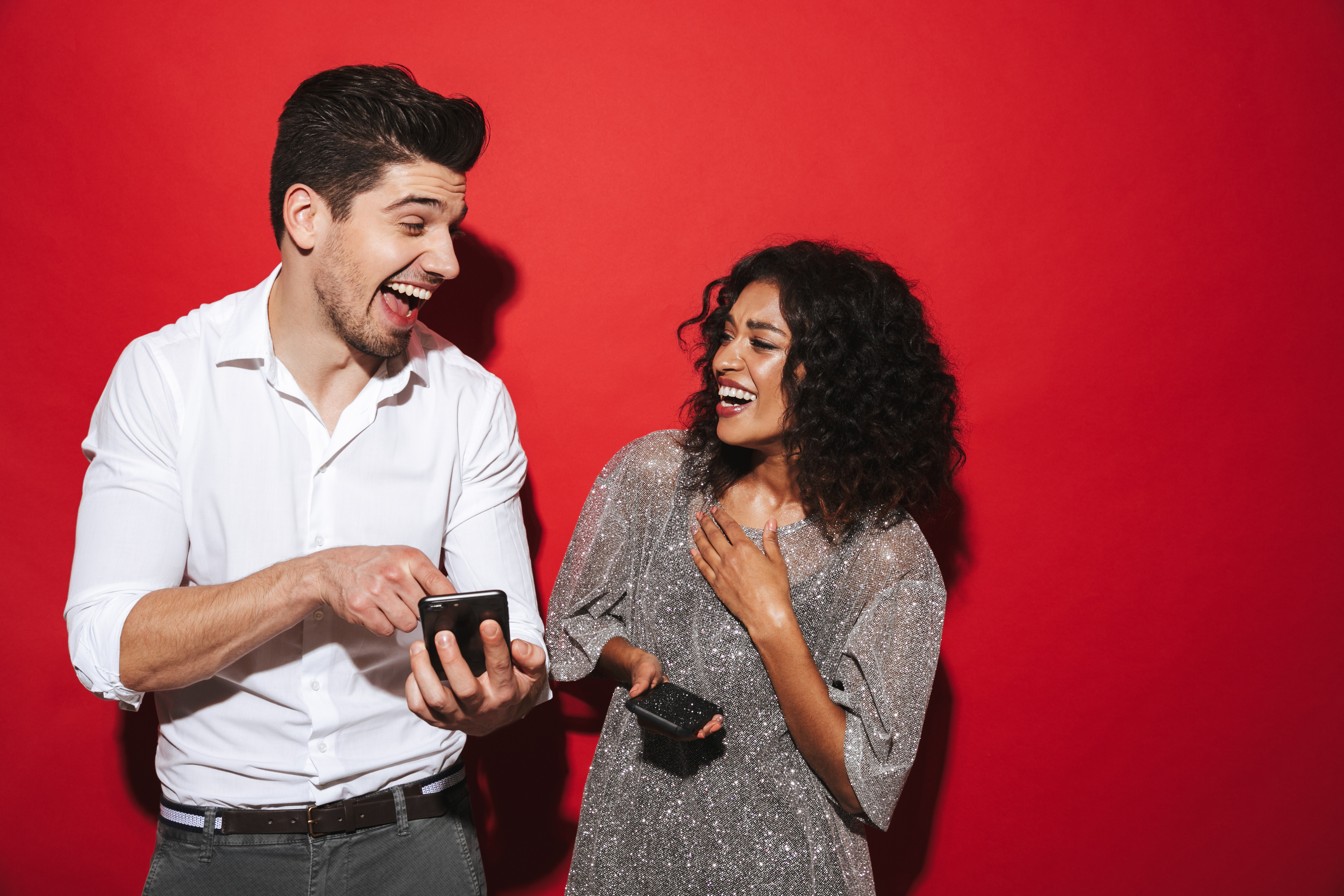 A man and woman laugh together as they point at their mobile phones in front of a red background.