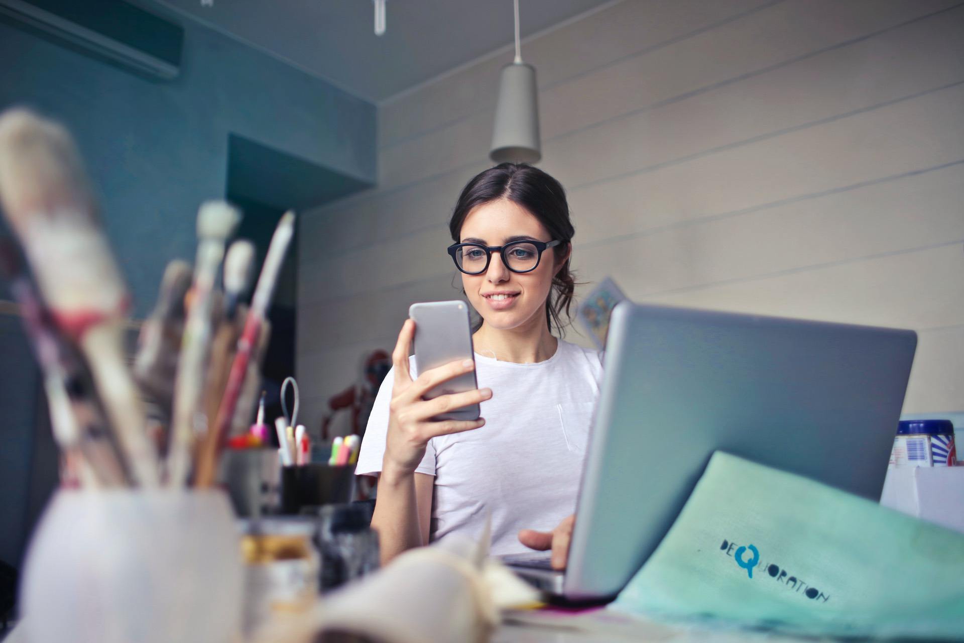 Woman in glasses sitting at a desk using a smartphone and laptop