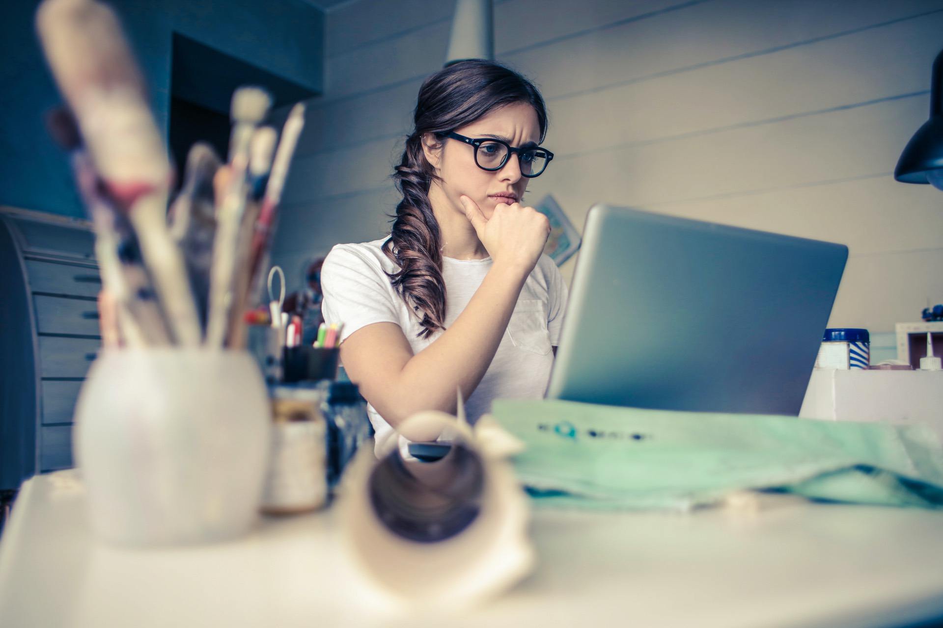Woman in glasses looks thoughtfully at a laptop in a creative workspace