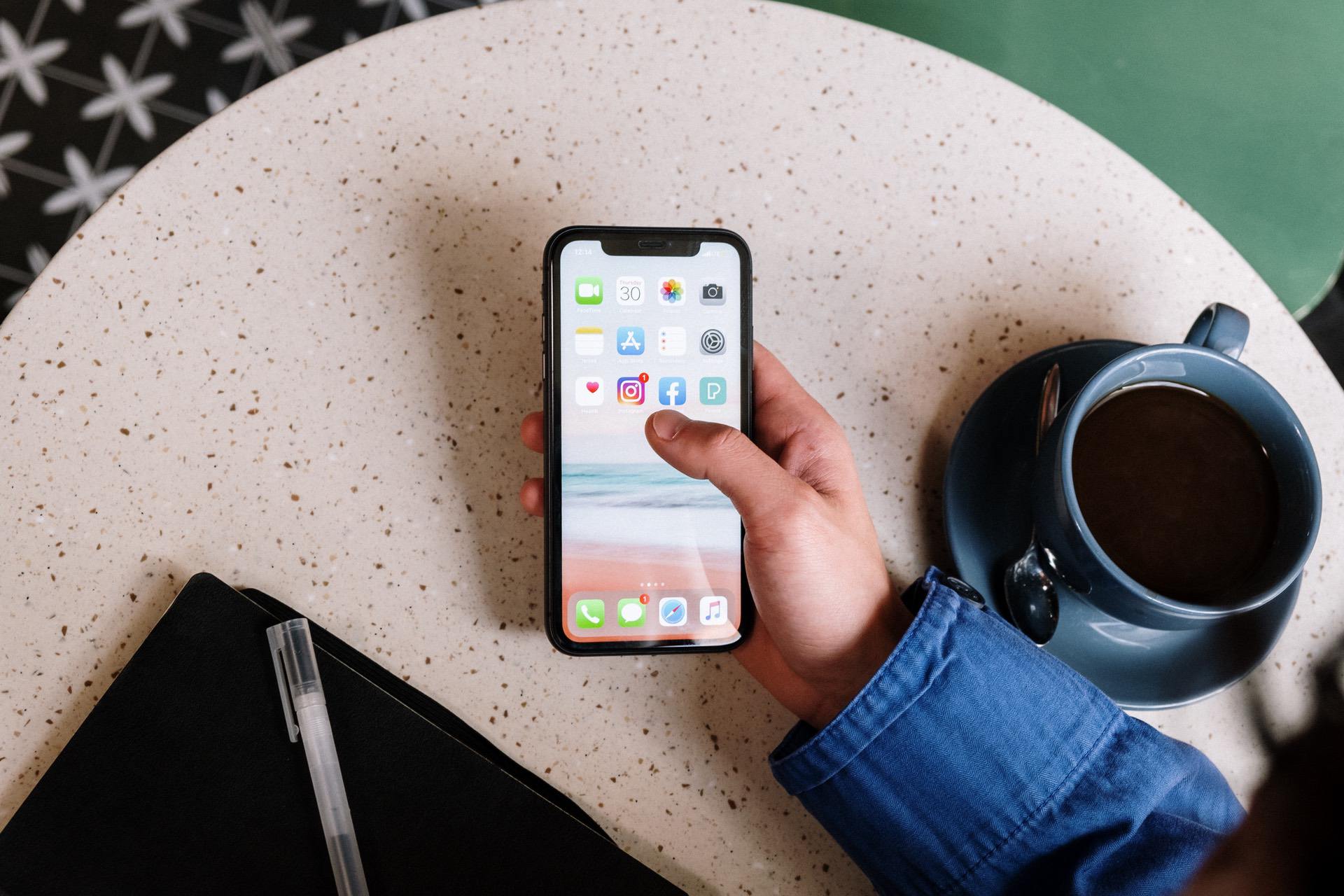 A person holding a smartphone with apps, coffee cup, and notebook on a table