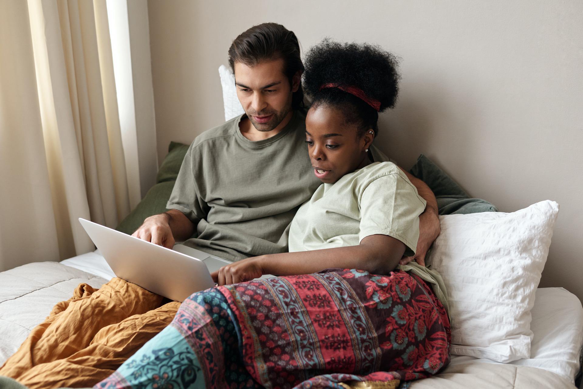 Couple seated on a bed, looking at a laptop together, sharing a relaxed moment