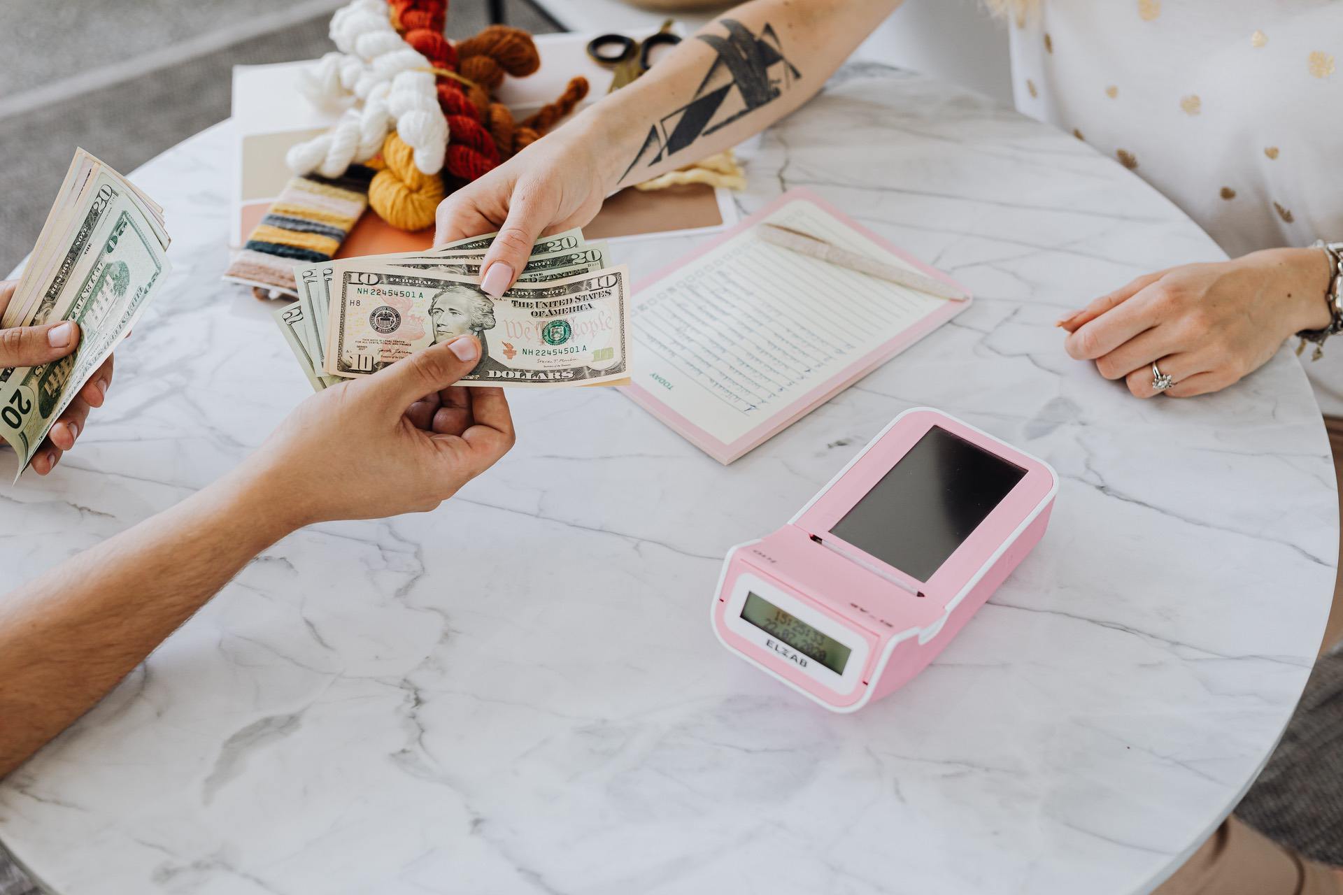 Hands exchanging cash over a marble table with a pink cash register in view