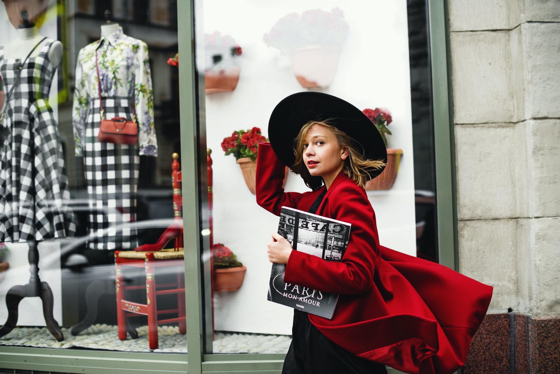 Woman in a red coat and black hat poses outside a boutique with fashion displays