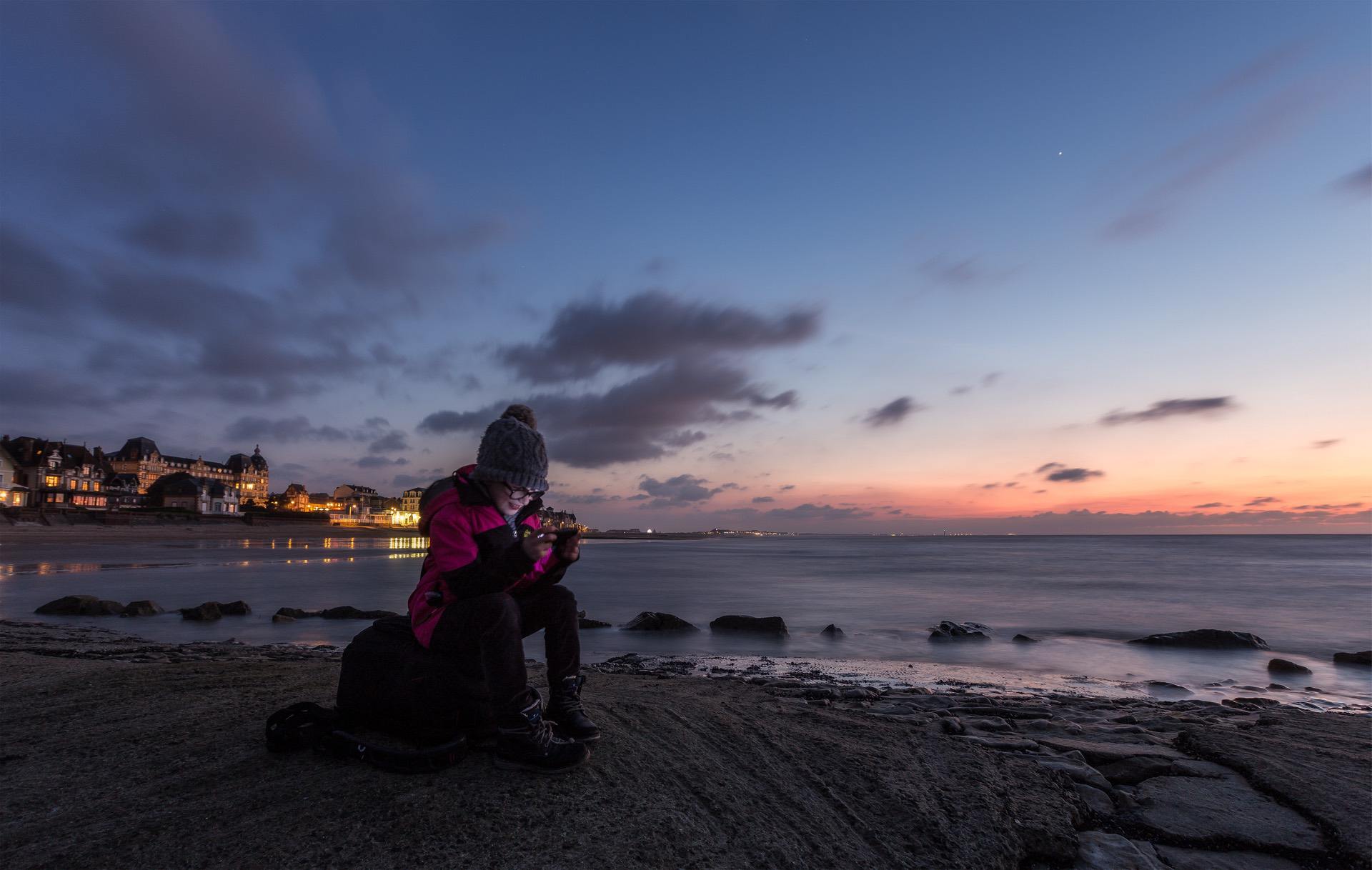 Person in winter clothing sitting on rocks by the beach at sunset, using a device